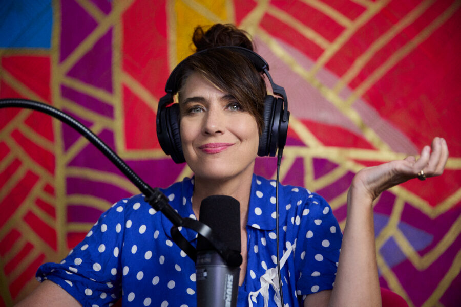 Kathleen Hanna smiling in front of a red, purple, and gold background while facing a microphone. She is wearing a blue shirt with white dots and black over-ear headphones.
