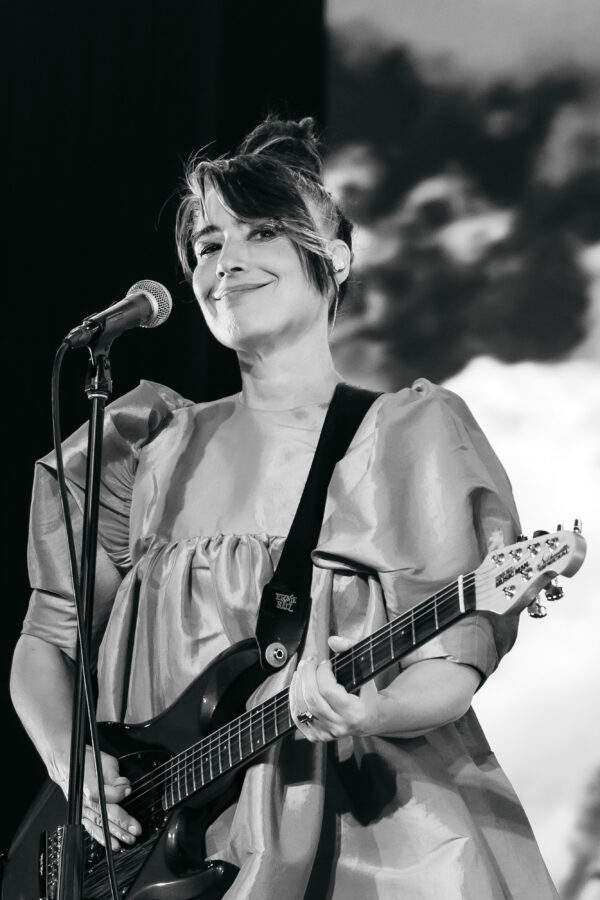 Black and white photo of an adult in front of a mic stand, smiling while holding an electric guitar