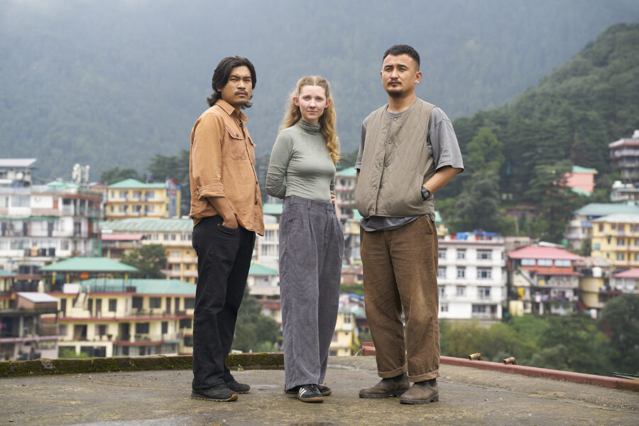 a woman stands between two men on a rooftop, with a city and greenery in the background