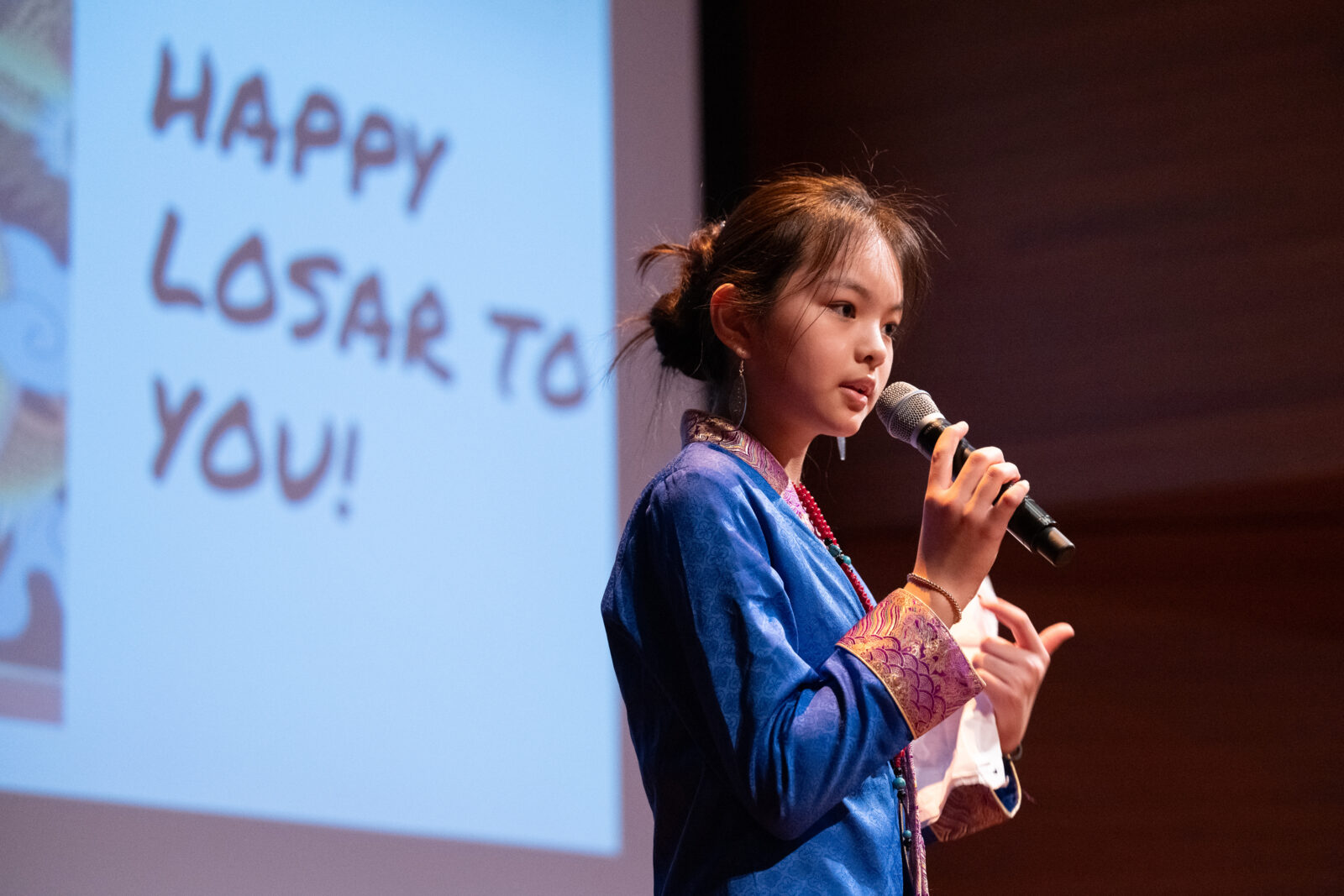 Child stands on stage with a microphone and behind her is a sign that reads, "Happy Losar to You!"