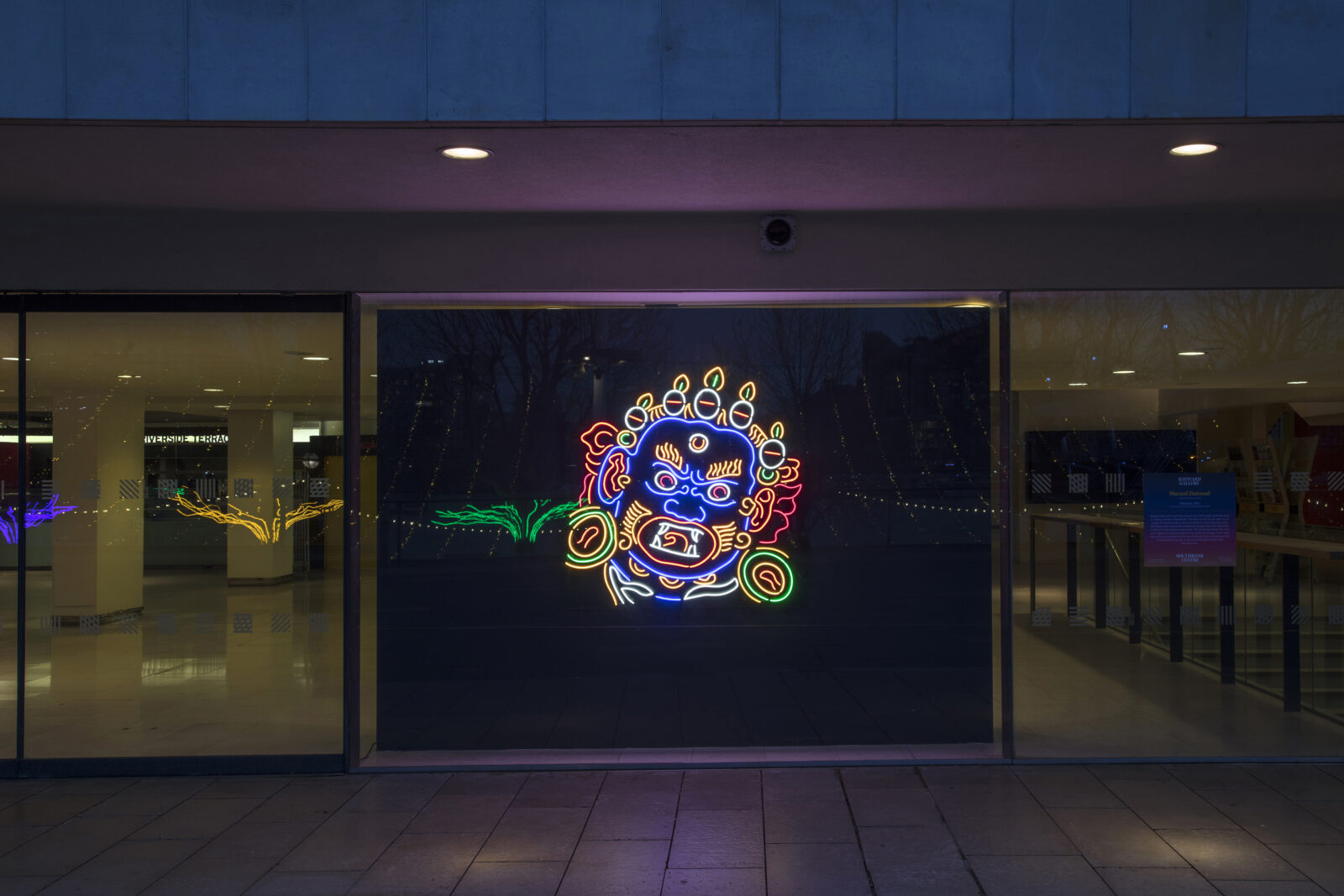 neon sculpture of Mahakala lit up on a black wall, seen from the outside of a building