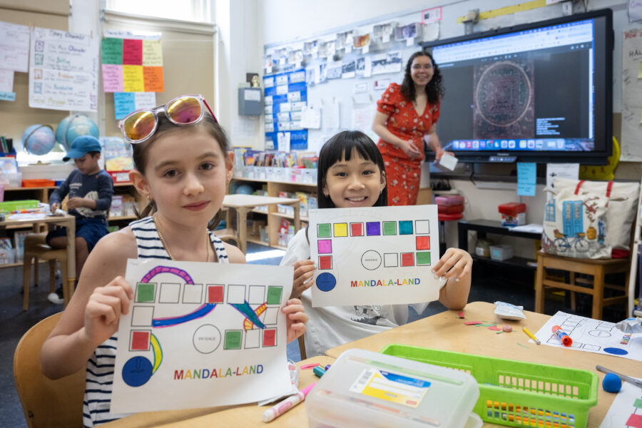 Two students in a classroom hold up a paper labeled "MANDALA-LAND" with a colorful grid of squares starting from circle labeled "ANGER" to a circle labeled "PATIENCE"
