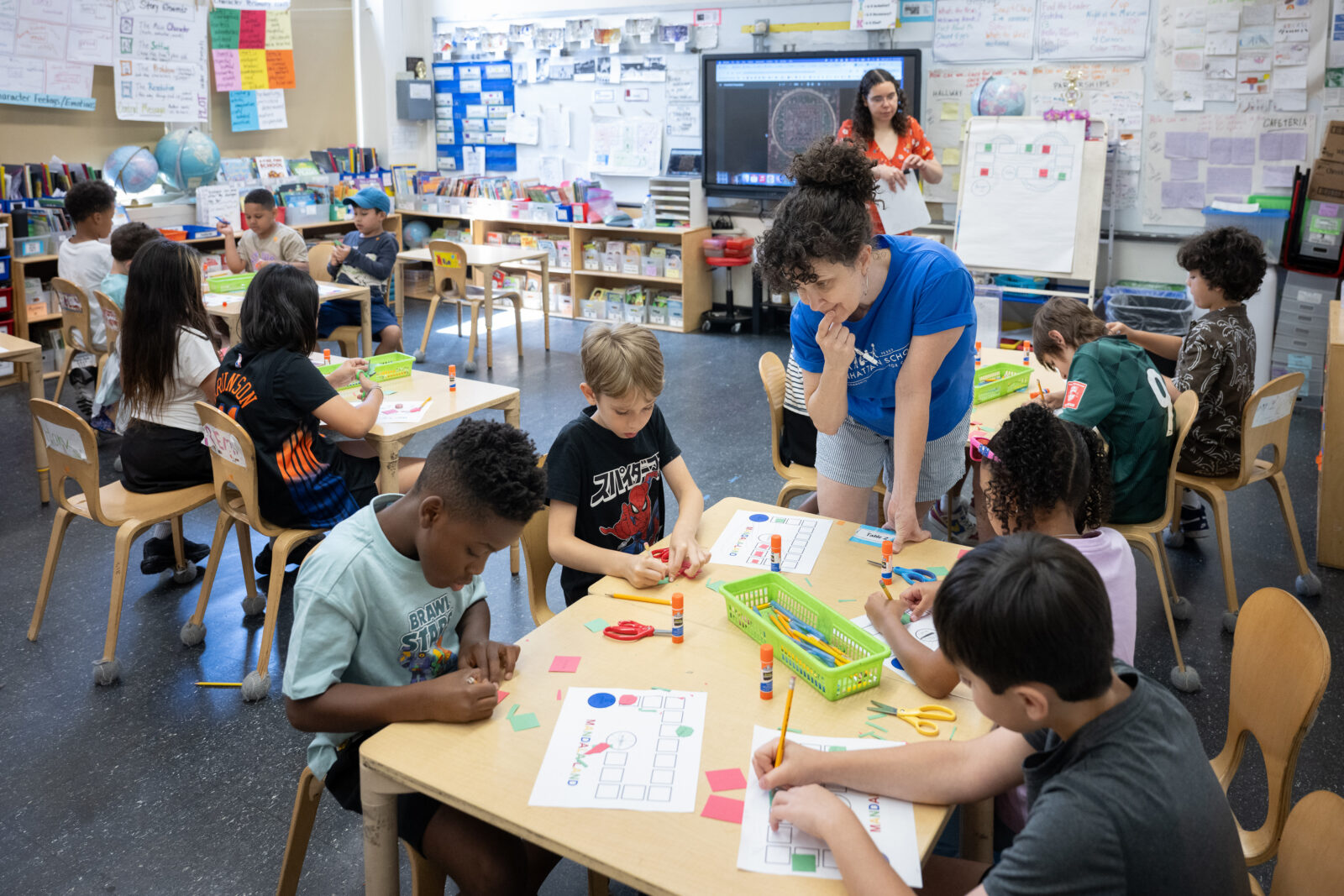 A teacher looks on as a group of students glue colorful pieces of paper onto a sheet of white paper