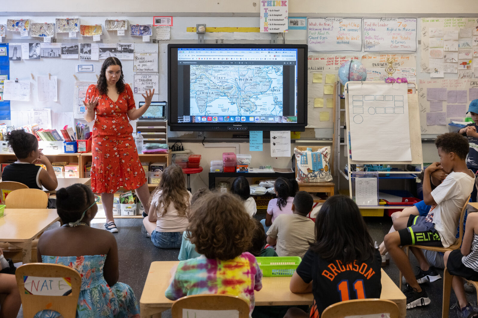 A teacher speaks in front of a group of students, standing beside a screen displaying a map of Asia