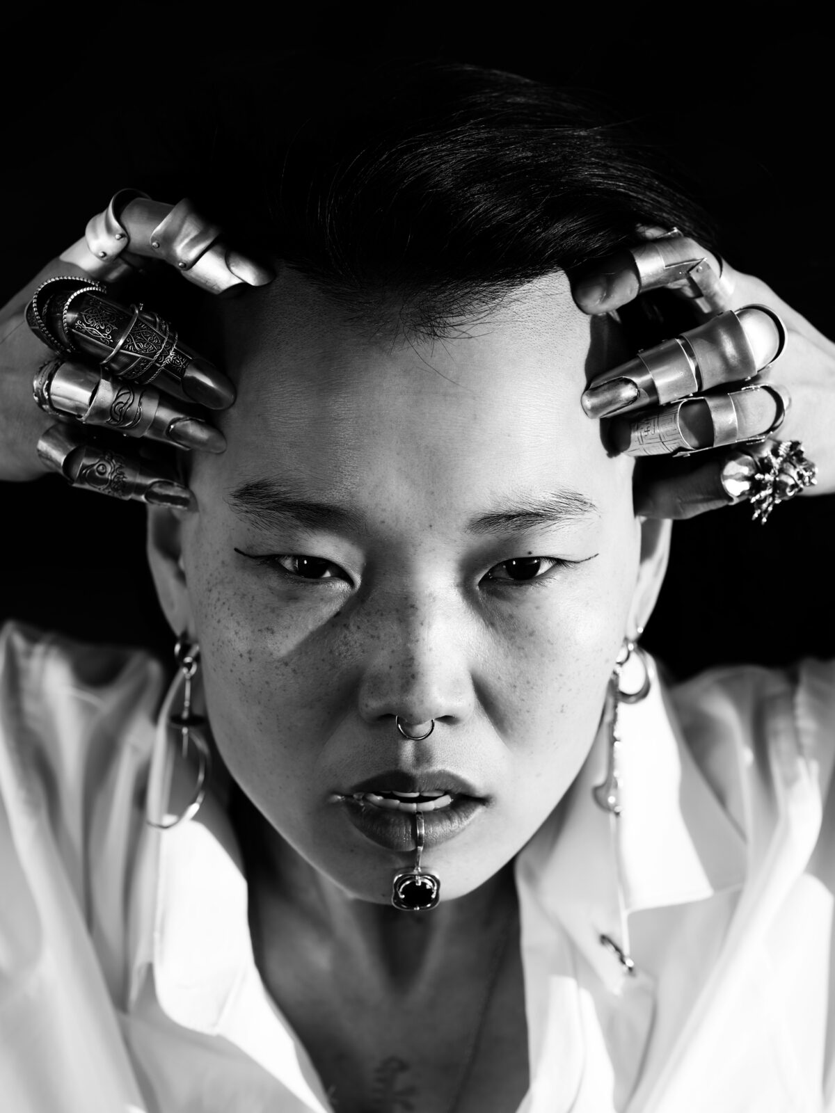 Black and white close up photo of an adult holding hands up to the top of their forehead, with rings on each finger