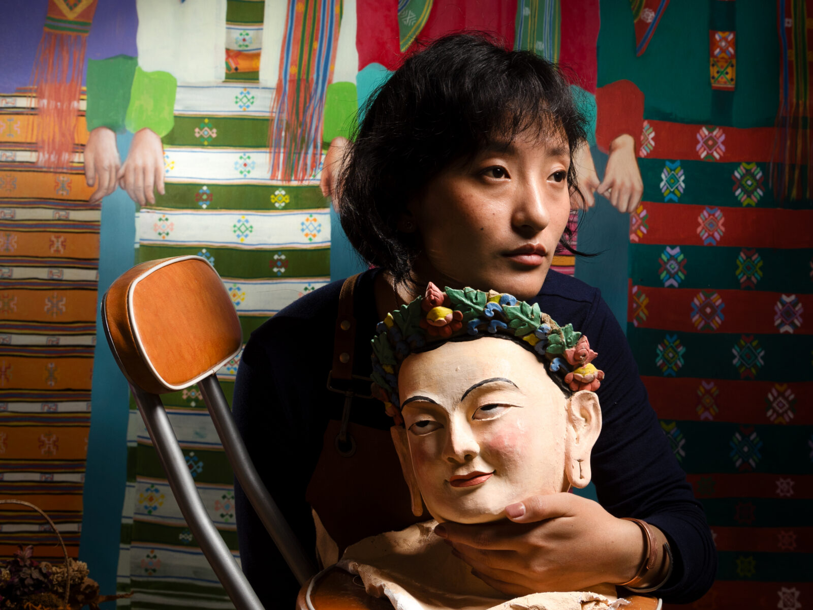 An adult with short black hair wearing a black sweater holds a sculpture of a head, while leaning on a leather stool