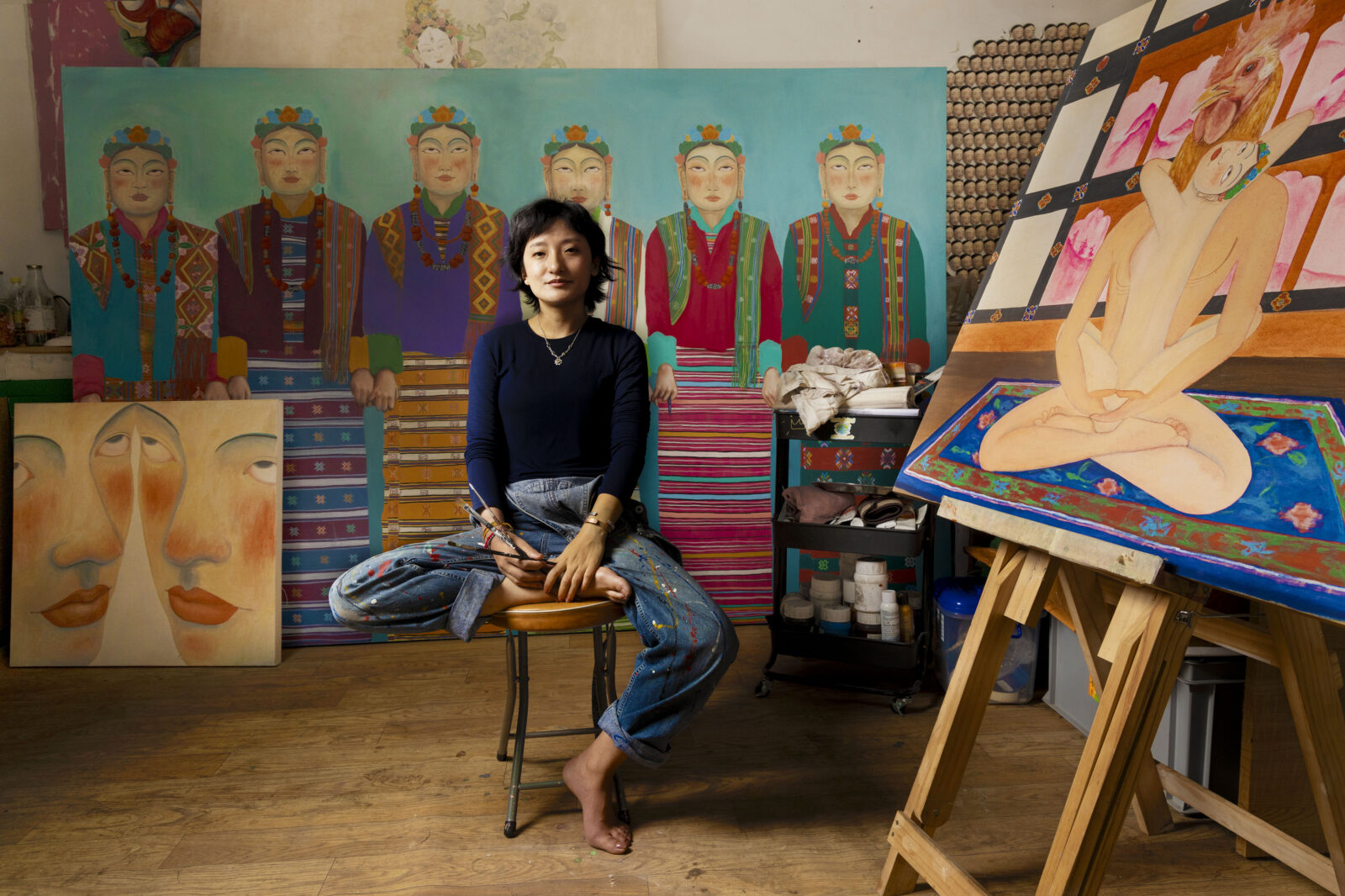 Artist Pem Lham sits cross-legged and barefoot on a stool in her studio in jeans and a dark long-sleeve top, holding paintbrushes in her lap. Behind her is a large painting of figures in traditional dress on a teal background, to her left is a close-up double-faced portrait, and to her right, on an easel, a colorful painting of a seated nude figure on a patterned rug.