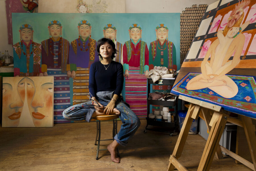 Artist Pem Lham sits cross-legged and barefoot on a stool in her studio in jeans and a dark long-sleeve top, holding paintbrushes in her lap. Behind her is a large painting of figures in traditional dress on a teal background, to her left is a close-up double-faced portrait, and to her right, on an easel, a colorful painting of a seated nude figure on a patterned rug.