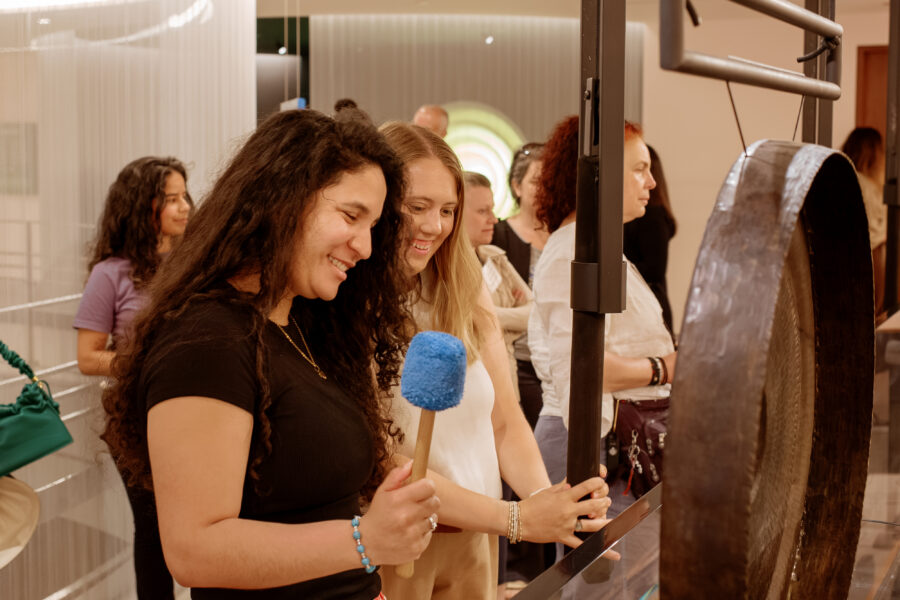 Two smiling women look at a gong, while one holds a mallet