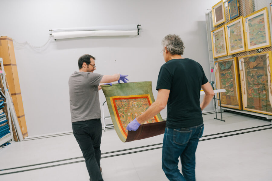 Two adult men each hold one end of a painting, carrying it toward a wall