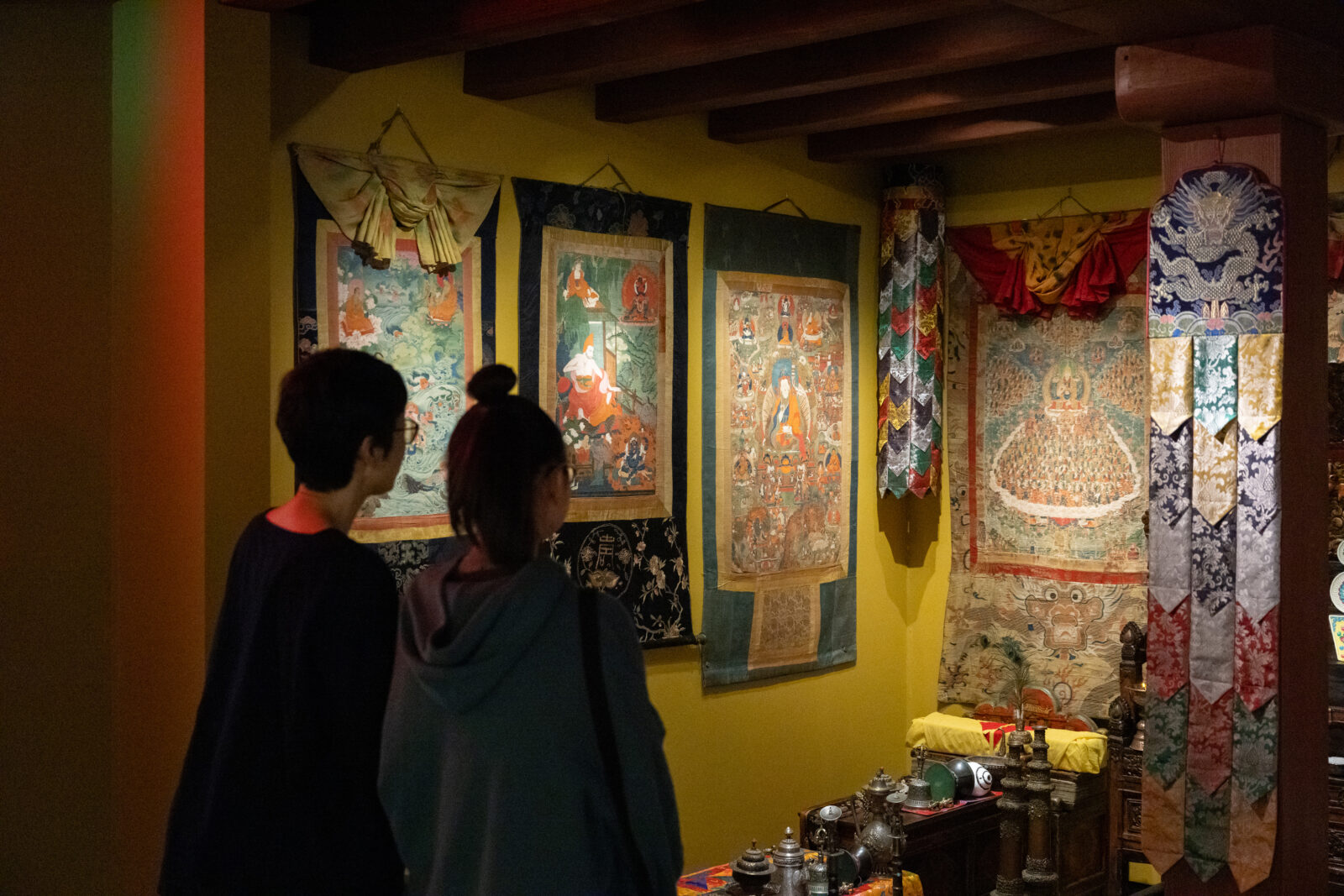 Two museum visitors look at a Tibetan Buddhist Shrine Room installation featuring scroll paintings, ritual implements, and colorful textiles.