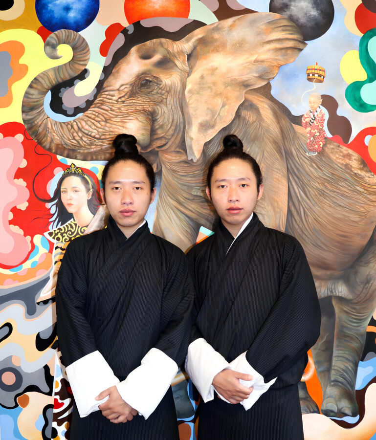 Two twins with matching top-knot hairstyles wearing black robes with white cuffs, stand side by side in front of a large, colorful mural of an elephant
