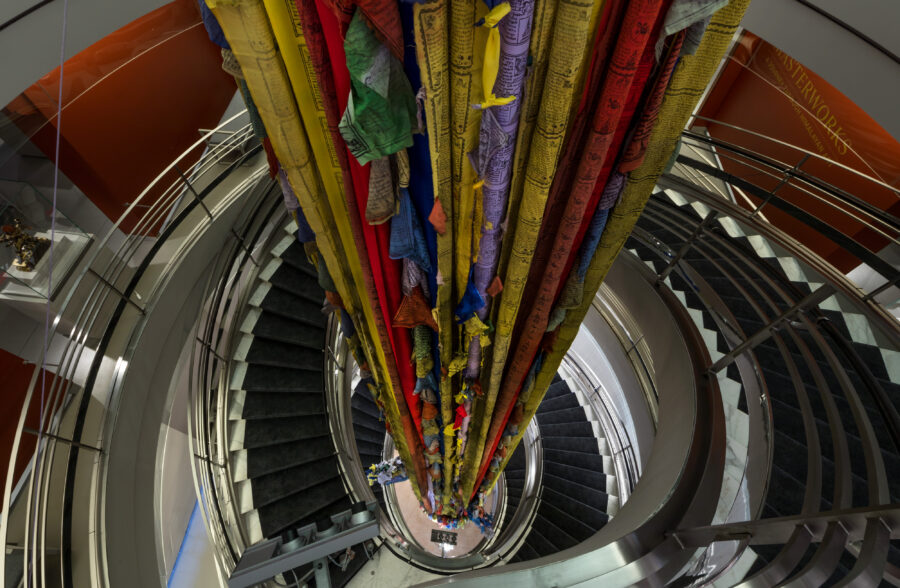 Photograph taken at the top of a spiral staircase looking down onto an artwork of prayer flags flowing down the center.