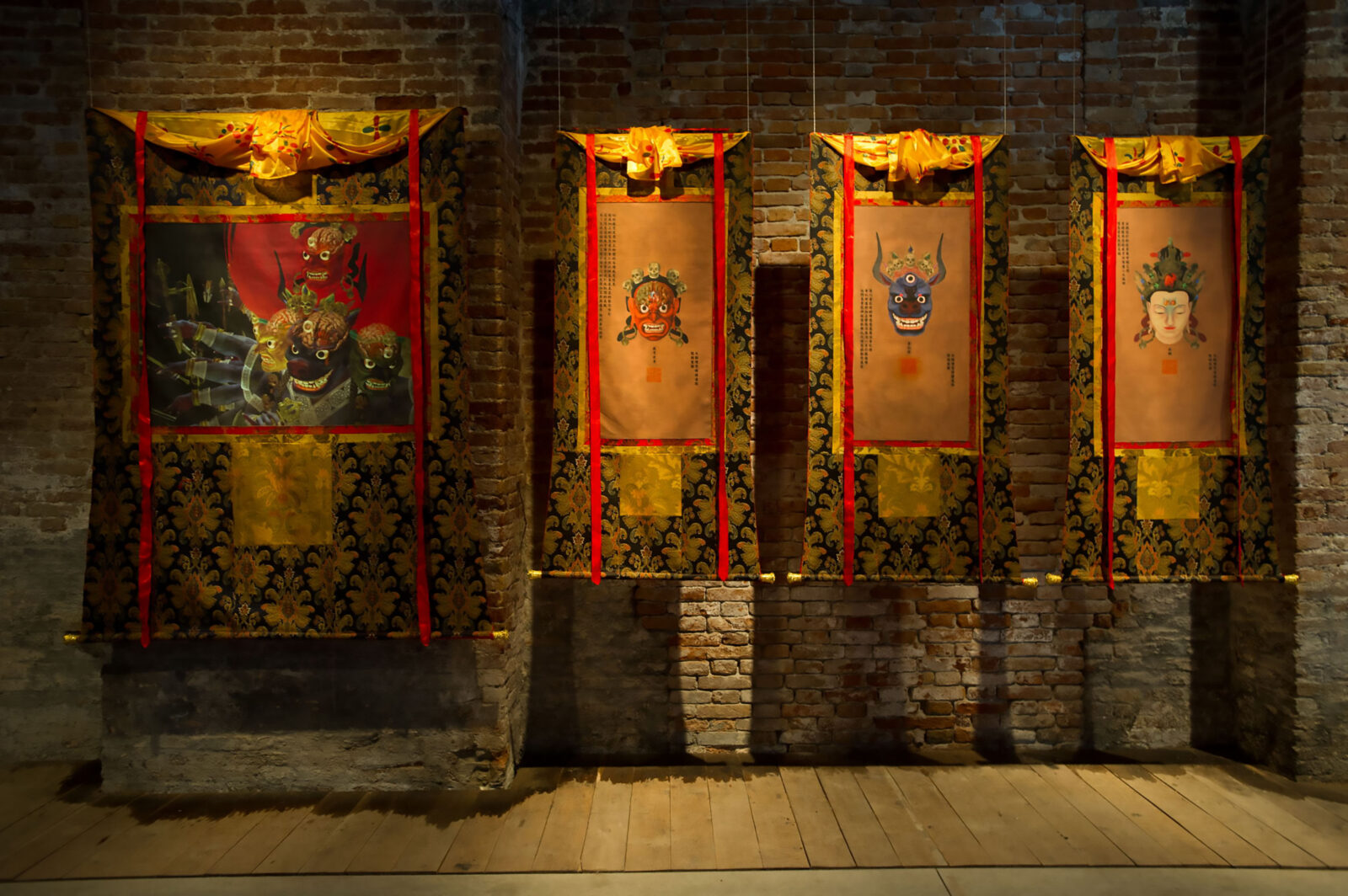 Four scroll paintings hanging in front of a brick wall, with one large painting to the left, and three slightly smaller and equal sized paintings to the right
